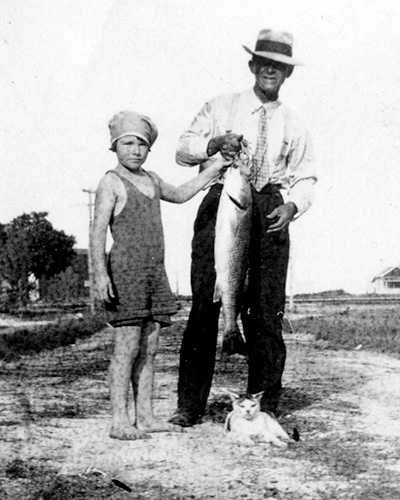 a young girl in her bathing suit and a man show off a fish they've caught as a cat lies at their feet